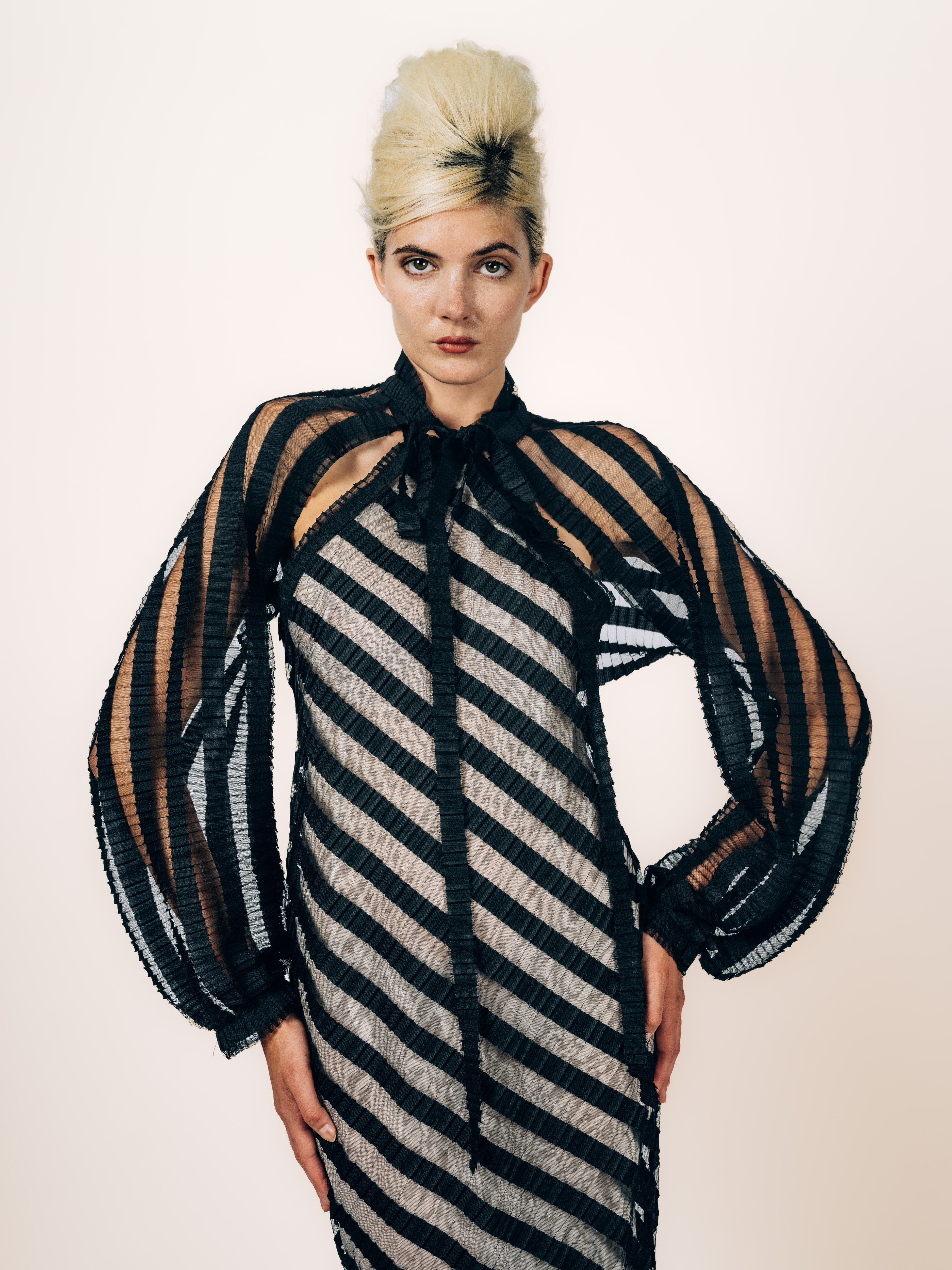 Transparentes Cape aus schwarzen Plissee-Streifen mit Ballonärmeln und Schleife, getragen über dem Neckholder-Abendkleid, von Maya Seyferth